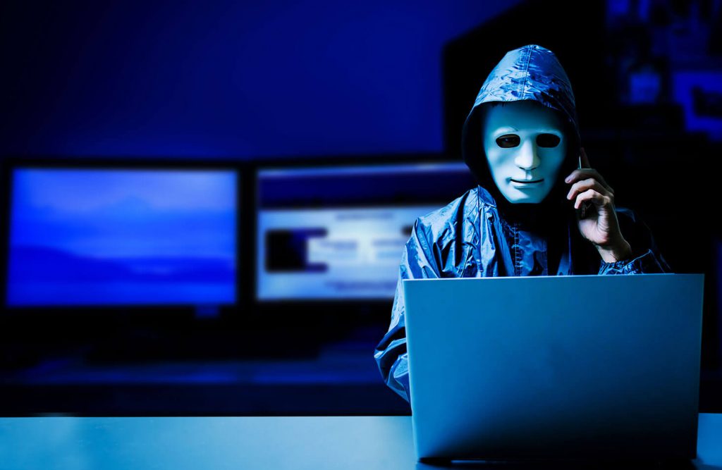 a person wearing a hooded jacket and with a sculpted mask over their face speaks on a phone while looking at a laptop in a dark room