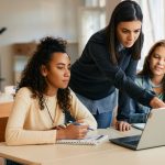 A woman stands between two female students pointing to a laptop screen.