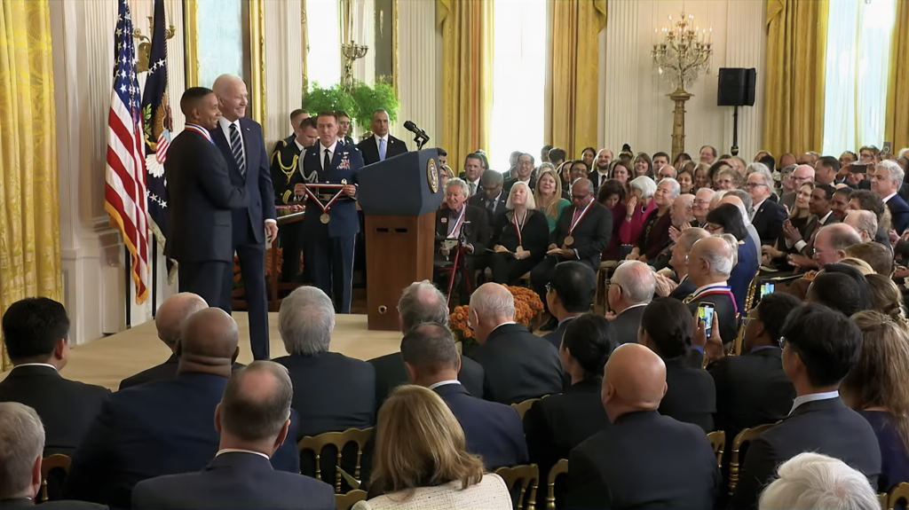 Image of Juan. Ε. Gilbert, PH.D., chair of the University of Florida Department of Computer & Information Science & Engineering, shaking hands with U.S. President Joe Biden in a room full of people attending a medal ceremony.